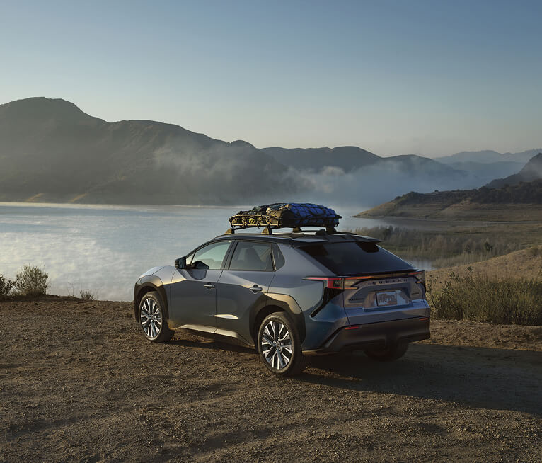 A 2026 Subaru Solterra electric SUV with a storage attachment on its roof-rails parked in front of a foggy lake in the sunlight.
